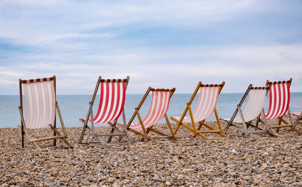 A landscape image of a pebble beach in the UK with 6 red and white stripey deck chairs lined up in a row